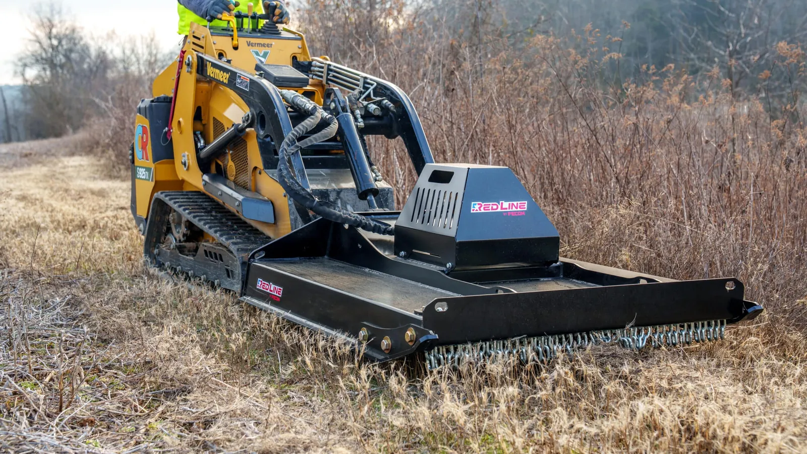 Stand-on mini skid steer with a rotary brush cutter mowing tall dry grass and brush